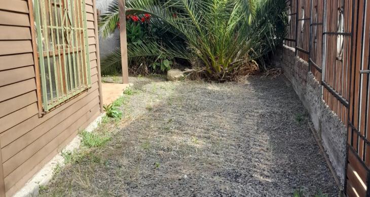 “Vista del antejardín lateral de la propiedad, donde se aprecia el portón de acceso principal construido en madera y metal.